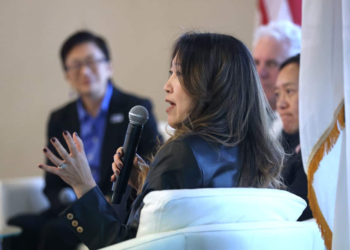 Panelist Julie Kim of Takeda is shown from the side speaking, gesturing with her left hand and speaking into a microphone, while the other panelists are seen in the background.