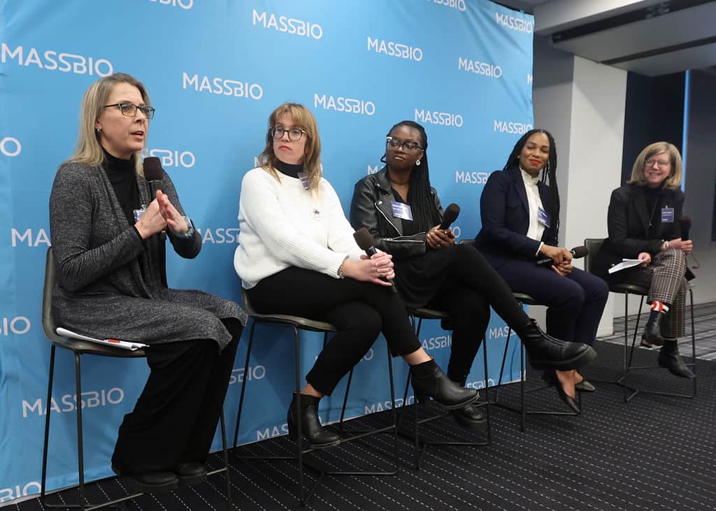 Four female panelists and a moderator hold microphones while sitting on high chairs in front of a MassBio branded step-and-repeat backdrop.