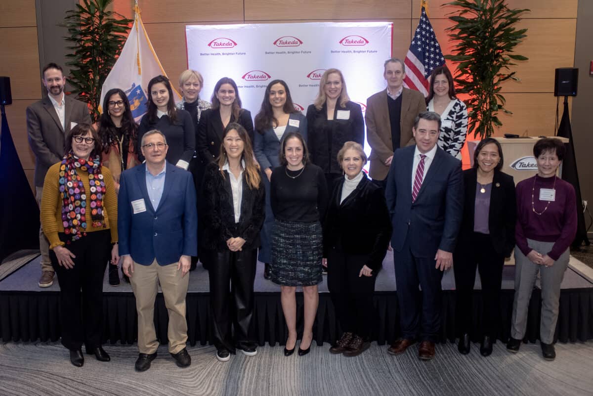 Standing in two rows in front of a Takeda banner, 17 of the attendees including Secretary Yvonne Hao, Julie Kim, Massachusetts Life Sciences Center staff, program sponsors and the awardees smile for the camera 