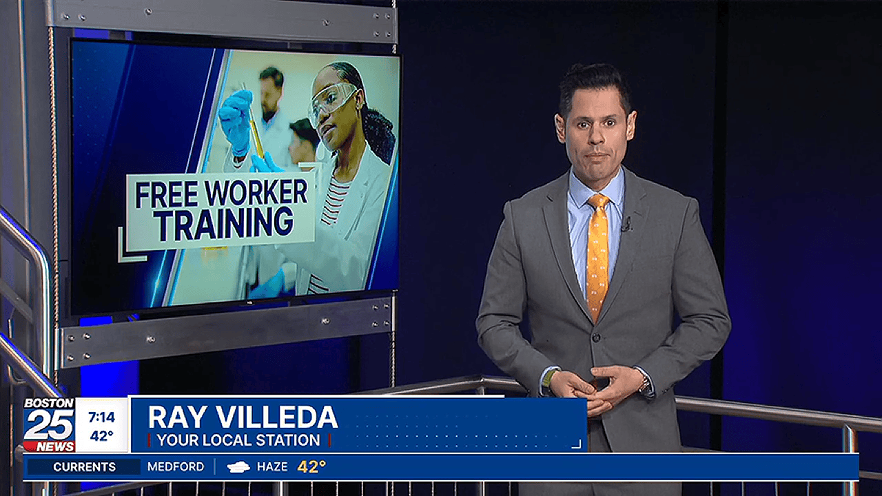 Ray Villeda of Boston 25 News stands in front of a large TV screen with the words "Free Worker Training" and a image of young people of color in a lab setting.
