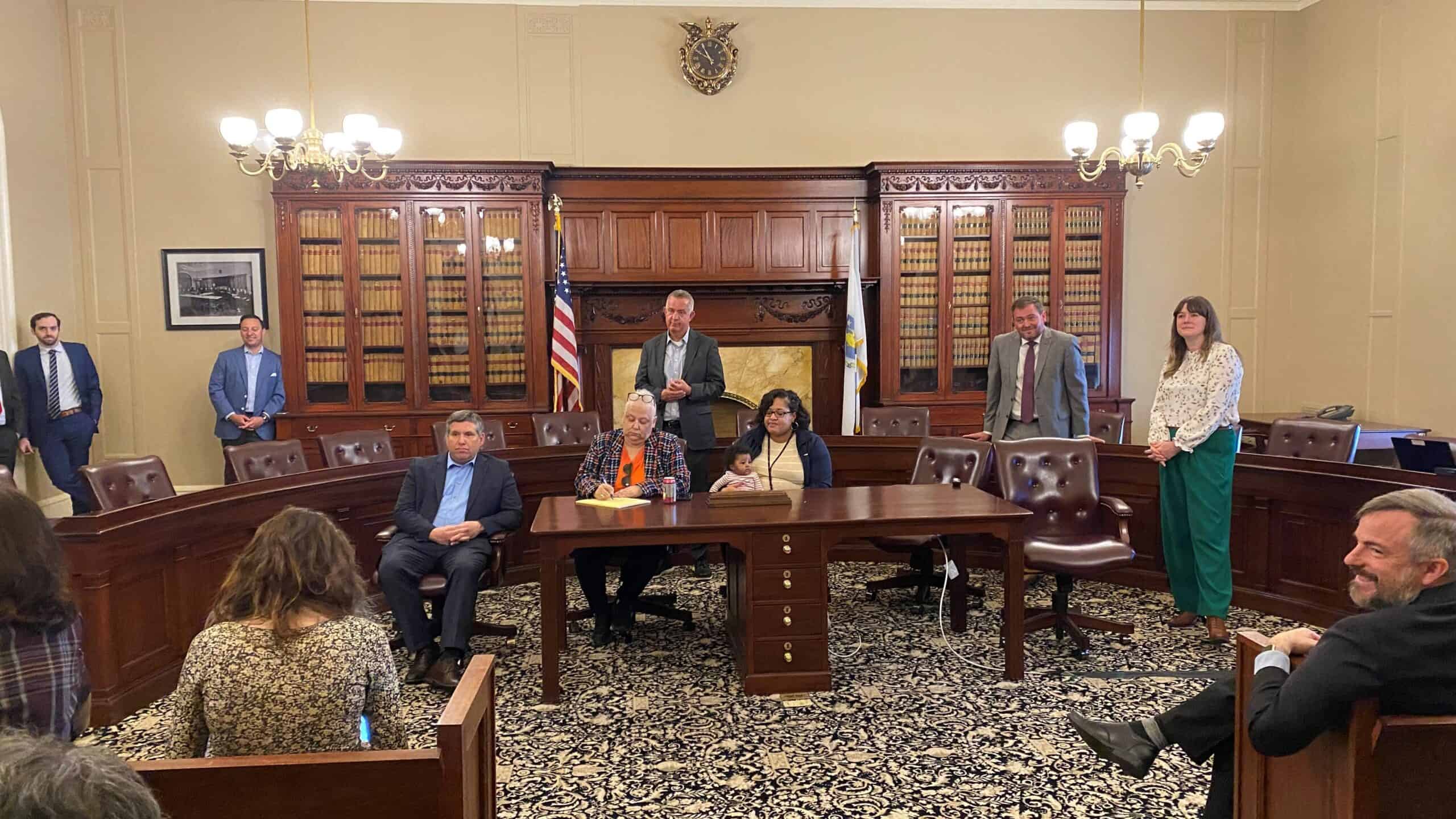 A group gathers in Room 222 at the Statehouse, and room with a large half circle desk with chairs behind it as well as a build-in bookcase that spans a large portion of the wall. There are individuals standing and seated in the photo, including legislators and officials from MassBio and MLSC. 