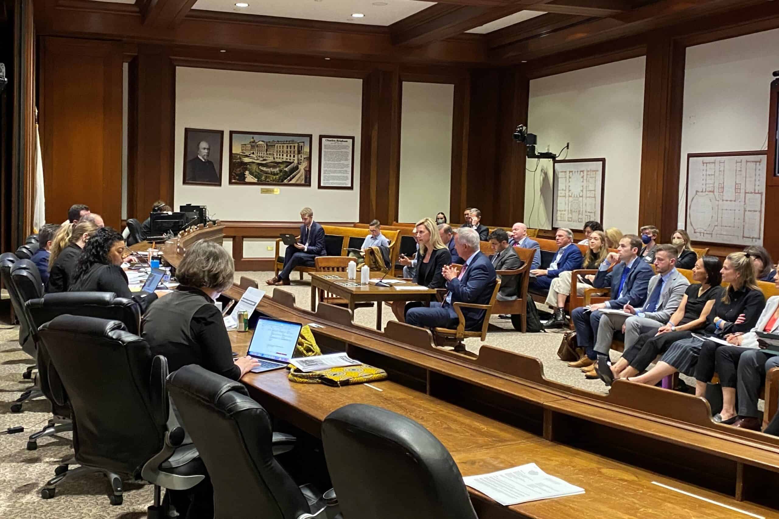 A hearing room in the Massachusetts State House with seats to the left for legislators and staff, a table in the middle for people testifying (in this case Kendalle Burlin O'Connell and Ed Coppinger), and then benches for guests to sit.