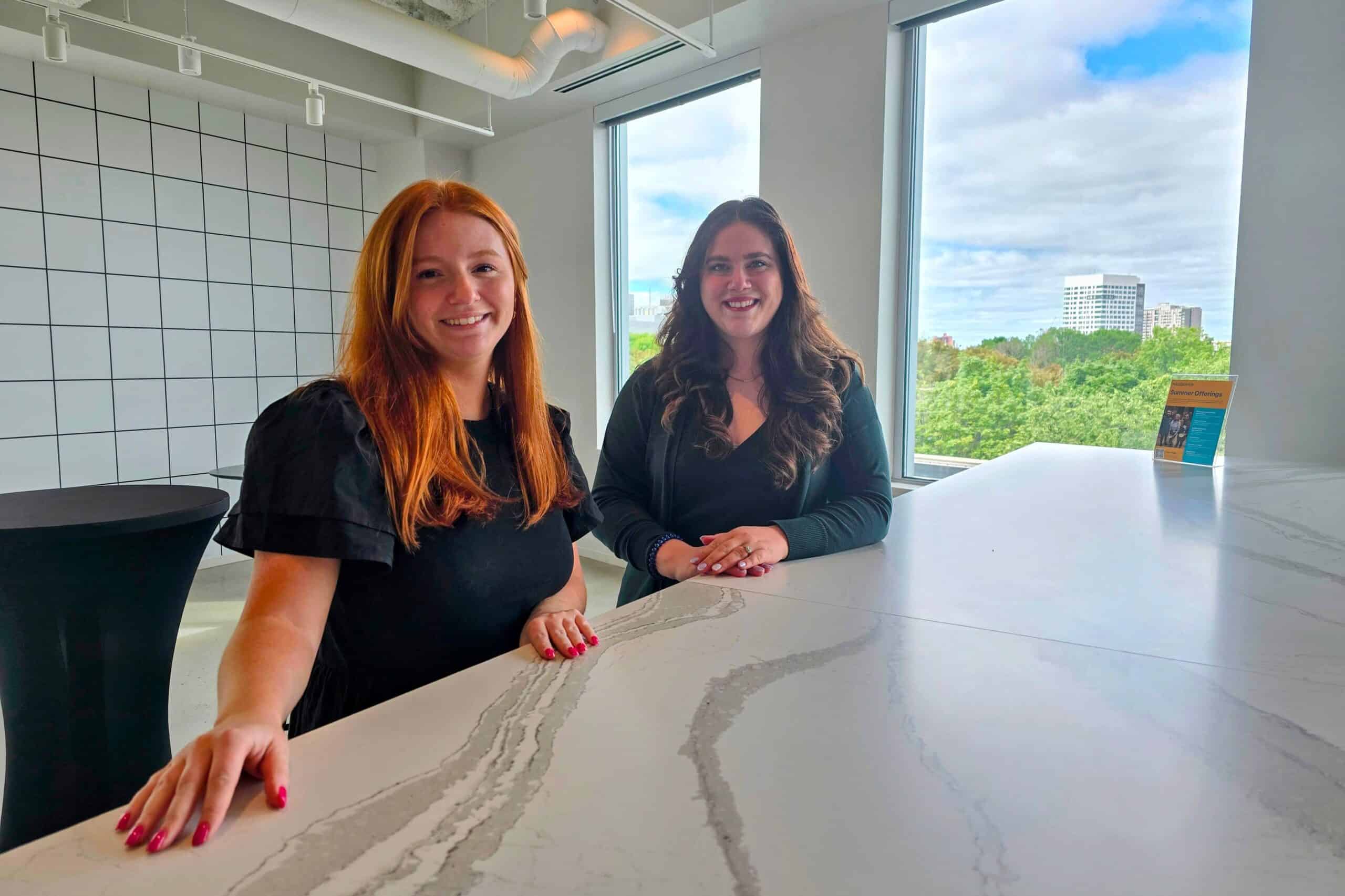 Two women behind a marble countertop with a window showing a lush tree landscape.