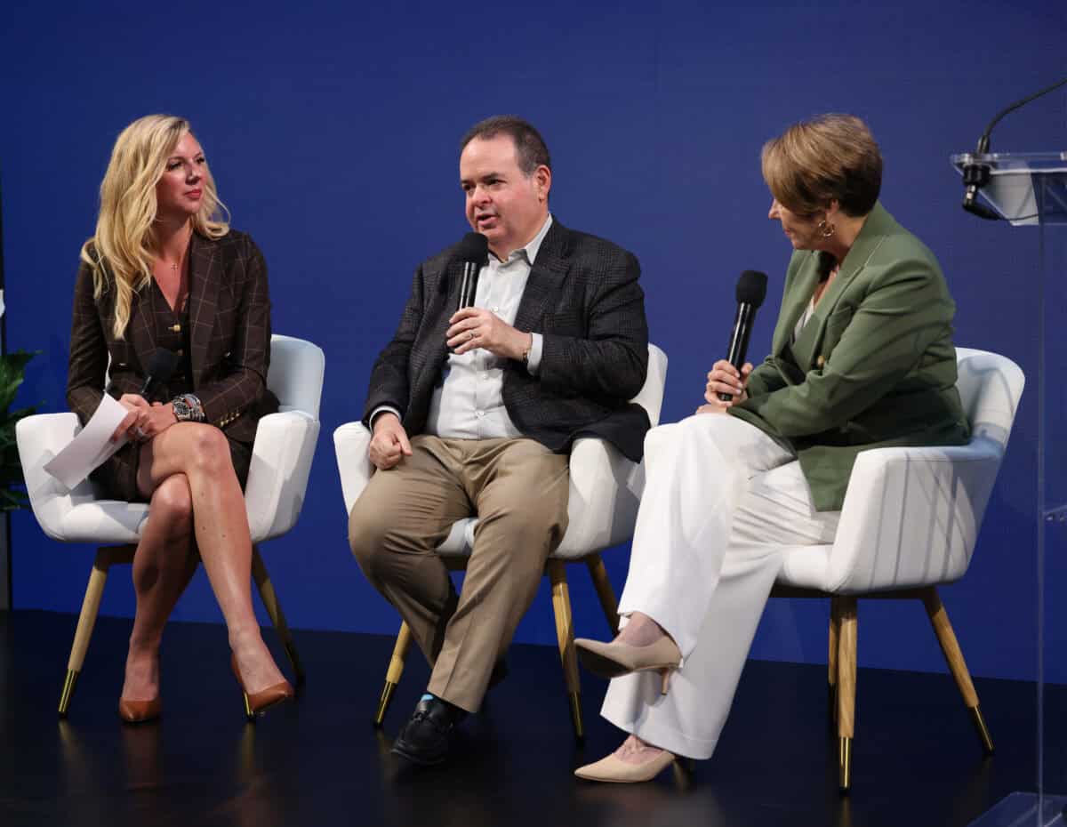 Three people sitting on a stage in white chairs each holding microphones. The woman on the left is wearing a skirt suit and has blonde hair; the man in the middle has khakis trousers, an opened colored shirt, and a blazer; and the woman on the right has a green blazer with white pants.