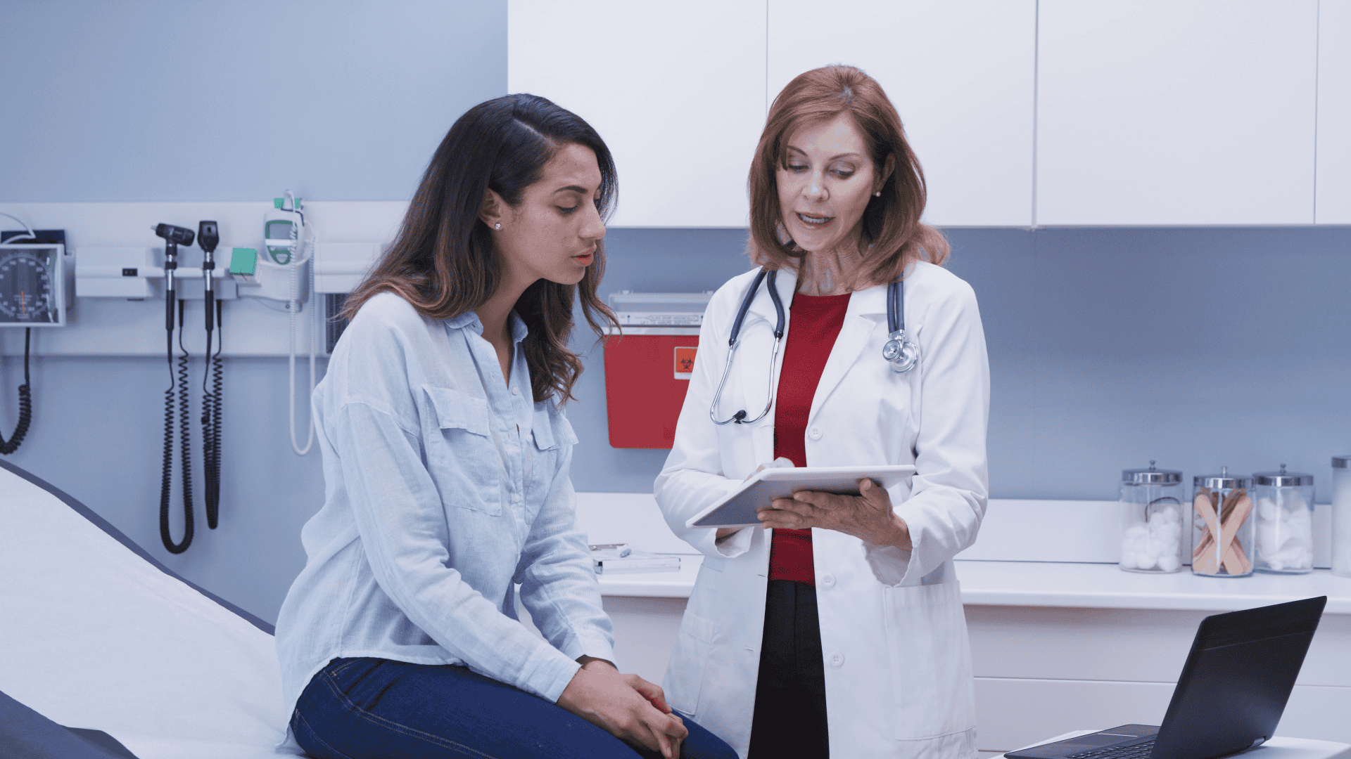 A female doctor with red hair is speaking with a younger female patient with dark hair while gesturing at a tablet in a medical examination room.